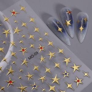 May include: Sheet of gold star nail art stickers with red and blue accents. The stickers are shown next to three long, oval-shaped nails with a blue and silver gradient design and gold star accents. The sheet is labeled "MO-09".