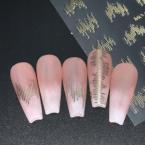May include: Five long, pink artificial nails with gold line designs. The nails are arranged on a black surface, with a sheet of gold nail art stickers in the background. The nail designs include vertical and horizontal lines.