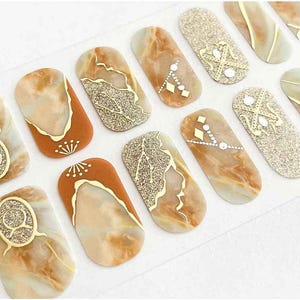 May include: A sheet of nail art stickers featuring various designs. Some stickers have a marble effect with gold accents, while others have glitter or jewel embellishments. The stickers are in shades of brown, gold, and white.