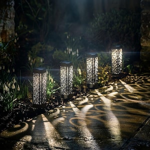 Può includere: Quattro luci solari rettangolari da giardino con intricati motivi ritagliati disposte lungo un sentiero in pietra. Ogni luce emette un bagliore bianco brillante, proiettando ombre decorative e vorticose sul terreno di notte.