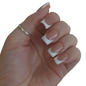 Puede incluir: Una mano con un anillo plateado en el dedo índice, mostrando uñas artificiales con punta francesa. Las uñas tienen una base beige natural con puntas blancas y forma cuadrada.