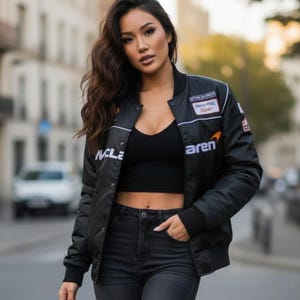 May include: A black bomber jacket featuring white McLaren branding, a small orange logo, and various racing-themed patches on the chest and sleeves. The jacket is worn open over a black ribbed crop top and dark gray denim jeans.