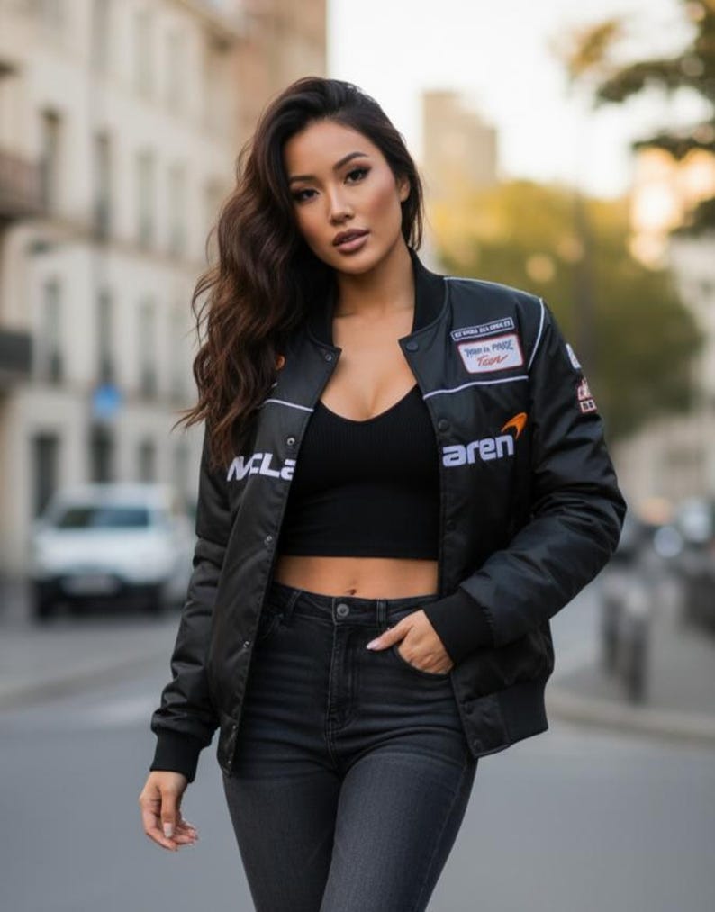 May include: A black bomber jacket featuring white McLaren branding, a small orange logo, and various racing-themed patches on the chest and sleeves. The jacket is worn open over a black ribbed crop top and dark gray denim jeans.