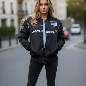 May include: A black satin-finish bomber jacket featuring the white McLaren logo across the chest, a white crew-neck t-shirt underneath, and black slim-fit trousers. The jacket includes various racing-themed patches on the upper chest area.