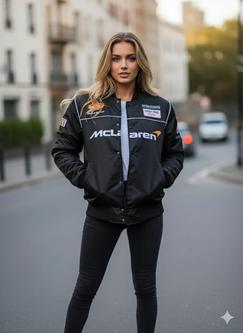 May include: A black satin-finish bomber jacket featuring the white McLaren logo across the chest, a white crew-neck t-shirt underneath, and black slim-fit trousers. The jacket includes various racing-themed patches on the upper chest area.