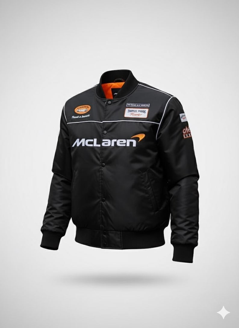 May include: A black bomber jacket featuring the McLaren logo in white and orange on the chest. The jacket includes a snap-button front closure, ribbed cuffs and hem, and various sponsor patches on the chest and sleeves with a bright orange interior lining.
