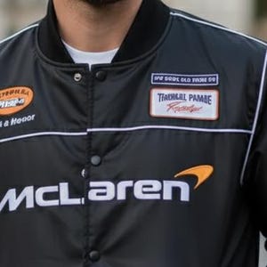 May include: A black button-up bomber jacket featuring the white McLaren logo on the chest. The jacket includes various embroidered patches, including an orange oval patch with text, a rectangular patch with text, and white piping along the shoulders.