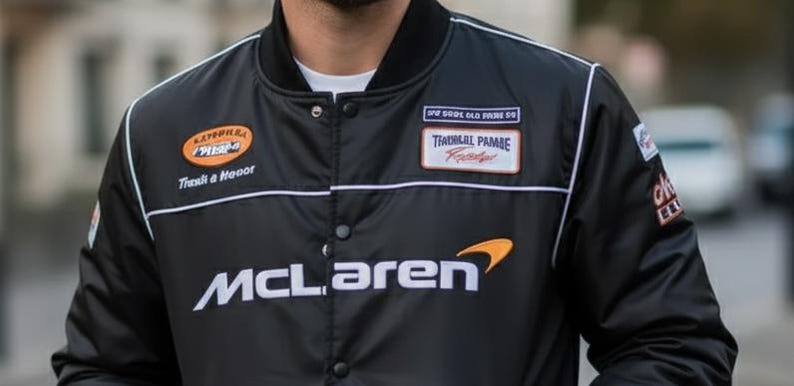 May include: A black button-up bomber jacket featuring the white McLaren logo on the chest. The jacket includes various embroidered patches, including an orange oval patch with text, a rectangular patch with text, and white piping along the shoulders.