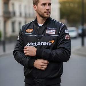 May include: A black satin bomber jacket featuring the McLaren logo in white and orange on the chest. The jacket includes a ribbed collar, cuffs, and hem, with various embroidered patches on the chest and sleeves, and a snap-button front closure.