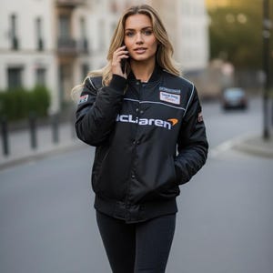 May include: A black bomber jacket featuring the text McLaren on the chest, a white horizontal stripe, and various small rectangular patches on the shoulders and chest. The jacket has a snap-button front closure, ribbed cuffs, and a ribbed collar.