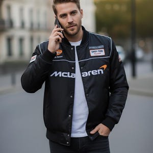 May include: A black bomber jacket featuring the white McLaren logo on the chest, a white t-shirt underneath, and dark denim jeans. The jacket includes various racing-style patches on the chest and sleeves, a snap-button closure, and ribbed cuffs.
