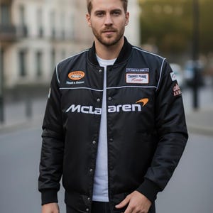 May include: A black bomber jacket featuring a white McLaren logo on the chest, a white crew neck t-shirt underneath, and various racing-style patches on the upper chest area. The jacket has a snap-button front closure and black ribbed cuffs.