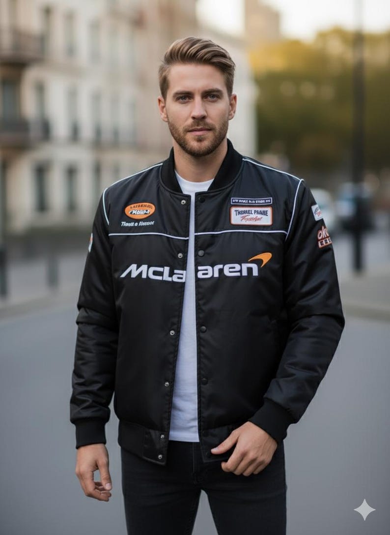 May include: A black bomber jacket featuring a white McLaren logo on the chest, a white crew neck t-shirt underneath, and various racing-style patches on the upper chest area. The jacket has a snap-button front closure and black ribbed cuffs.