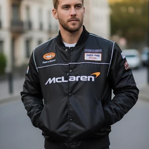 May include: A black bomber jacket featuring the McLaren logo in white and orange on the chest, with various racing-themed patches on the upper front, a snap-button closure, ribbed collar and cuffs, and white piping details along the shoulders.