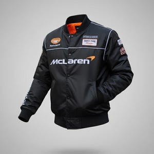 May include: A black bomber jacket featuring the McLaren logo in white and orange on the chest. The jacket has a snap-button front closure, a ribbed collar, cuffs, and hem, and an orange interior lining. Various racing-themed patches are visible.