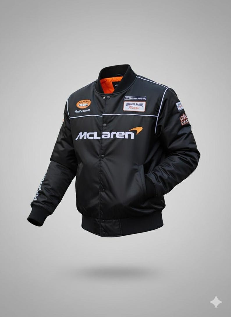 May include: A black bomber jacket featuring the McLaren logo in white and orange on the chest. The jacket has a snap-button front closure, a ribbed collar, cuffs, and hem, and an orange interior lining. Various racing-themed patches are visible.