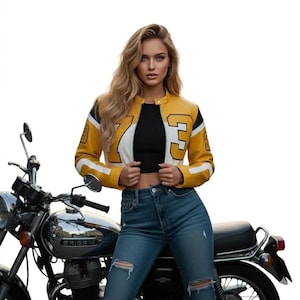 May include: A yellow, black, and white cropped leather jacket with the number 73 on the left sleeve and front. The jacket is paired with blue jeans and a black top. A motorcycle is in the background.