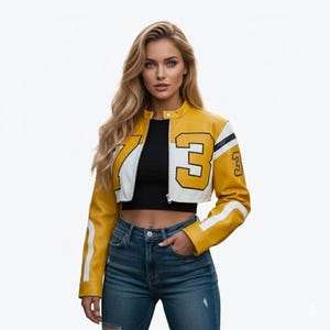 Può includere: Giacca corta gialla e bianca con il numero 73 a sinistra e il numero 3 a destra. La giacca ha un colletto nero e strisce bianche sulle maniche. La modella indossa jeans blu e un top nero.