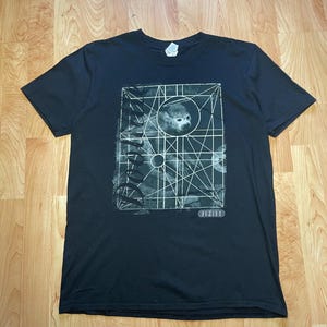 May include: Black t-shirt with a geometric design featuring a monkey's face within a circle. The design incorporates lines and shapes, with the word "FUTUR" at the bottom. The shirt is made of a soft material.