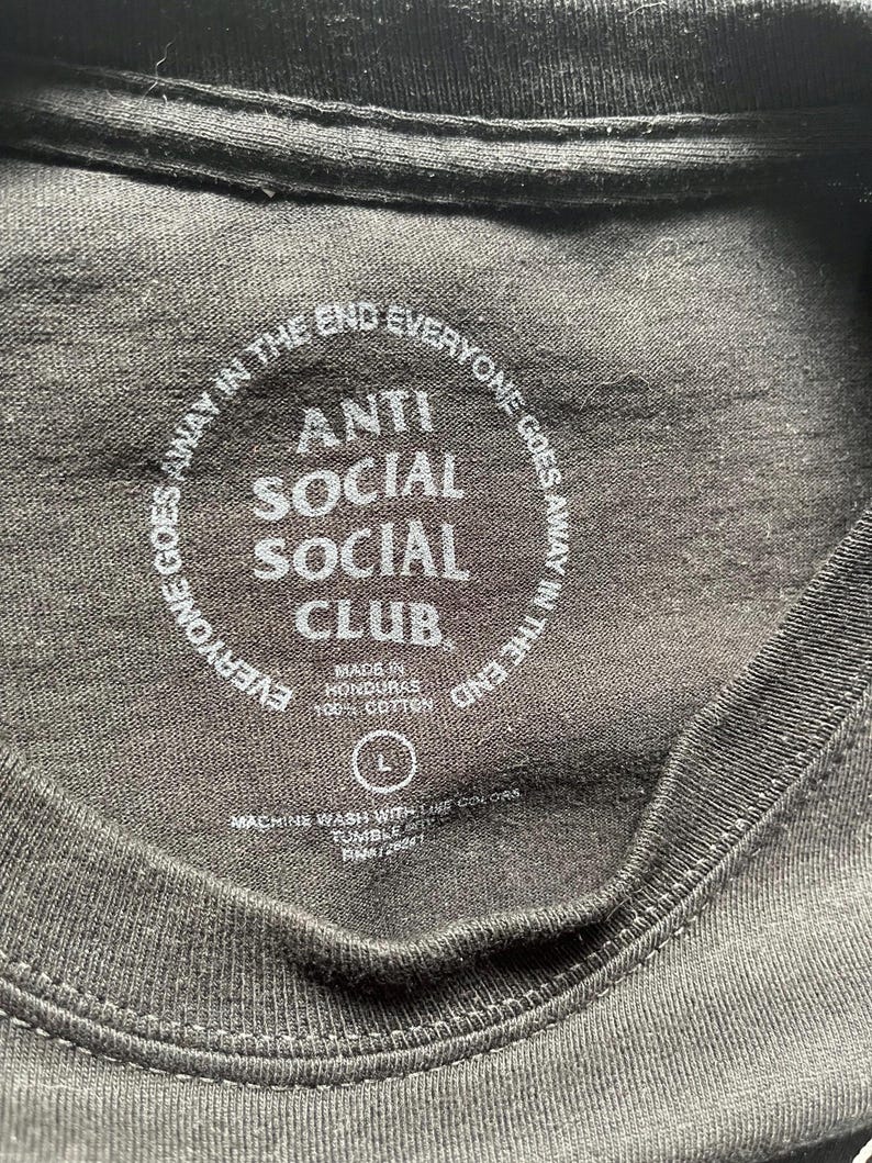 Puede incluir: Primer plano del cuello de una camiseta negra con el texto "ANTI SOCIAL SOCIAL CLUB" en blanco. El texto "EVERYONE GOES AWAY IN THE END" est&aacute; impreso en c&iacute;rculo alrededor del texto principal. La camiseta es talla L y est&aacute; hecha en Honduras.