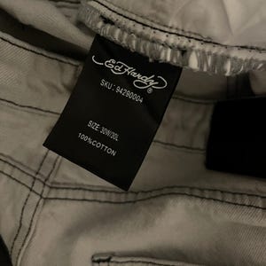 Puede incluir: Primer plano de unos vaqueros gris claro con costuras negras. Una etiqueta negra muestra el logotipo de Ed Hardy, el SKU, la talla 30W/30L y 100% algod&oacute;n. Los vaqueros est&aacute;n doblados, mostrando el forro interior.