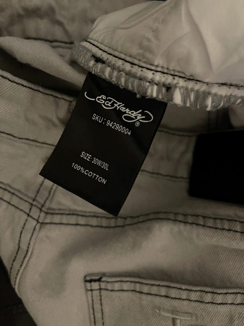 Puede incluir: Primer plano de unos vaqueros gris claro con costuras negras. Una etiqueta negra muestra el logotipo de Ed Hardy, el SKU, la talla 30W/30L y 100% algod&oacute;n. Los vaqueros est&aacute;n doblados, mostrando el forro interior.