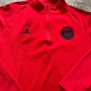 May include: A red athletic top with a quarter-zip closure and a mock neck. The top features a black Jumpman logo on the left chest and a Paris Saint-Germain team crest on the right chest. The long-sleeved top is made of a smooth, likely synthetic material.