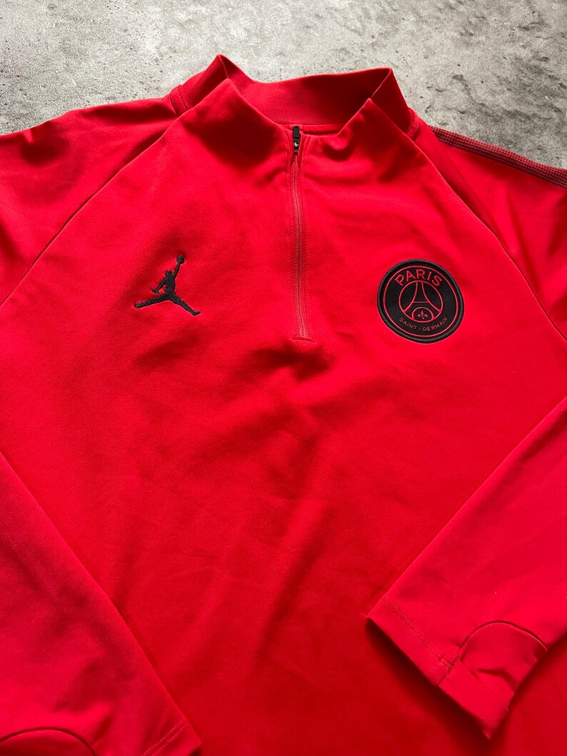 May include: A red athletic top with a quarter-zip closure and a mock neck. The top features a black Jumpman logo on the left chest and a Paris Saint-Germain team crest on the right chest. The long-sleeved top is made of a smooth, likely synthetic material.