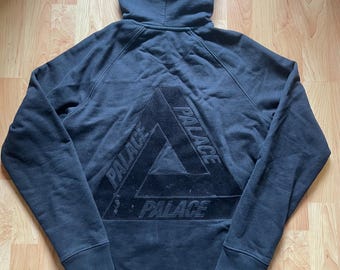 May include: Black hooded sweatshirt with the word "PALACE" in a triangle design on the back. The sweatshirt has a drawstring hood and ribbed cuffs and hem. The fabric appears to be a soft, comfortable material.