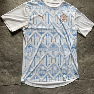 May include: White and light blue soccer jersey with a geometric pattern. The jersey features a Puma logo and a team crest. The sleeves are solid white. The jersey is made of a lightweight, breathable material.