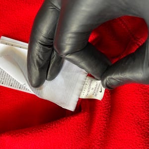 May include: Close-up of a red garment with a white tag. The tag has text and a Nike logo. The garment is being held by a gloved hand. The tag includes the size M and the code AQ2666-657.