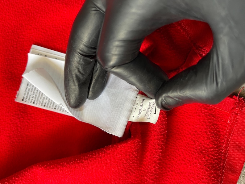 May include: Close-up of a red garment with a white tag. The tag has text and a Nike logo. The garment is being held by a gloved hand. The tag includes the size M and the code AQ2666-657.