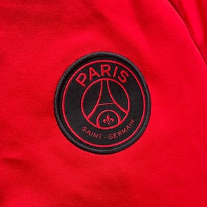 May include: Close-up of a red fabric with a circular black patch. The patch features the words "PARIS" and "SAINT-GERMAIN" in white, with a red outline. The design includes the Eiffel Tower and a fleur-de-lis.