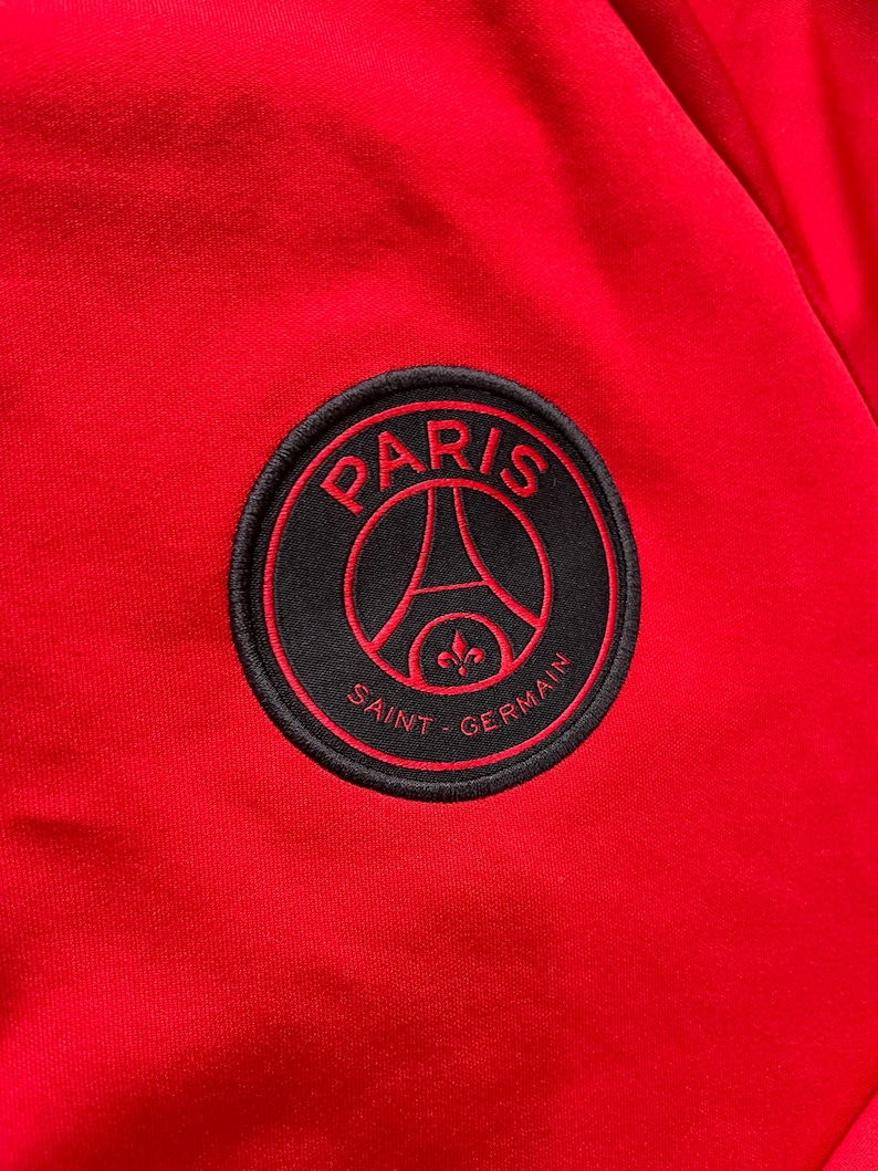 May include: Close-up of a red fabric with a circular black patch. The patch features the words "PARIS" and "SAINT-GERMAIN" in white, with a red outline. The design includes the Eiffel Tower and a fleur-de-lis.