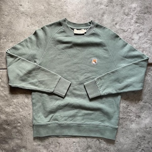 Puede incluir: Sudadera verde salvia con un pequeño logotipo de zorro en el pecho. La sudadera de cuello redondo tiene mangas largas con puños acanalados y dobladillo acanalado. La etiqueta de la marca es visible en la parte posterior del cuello.