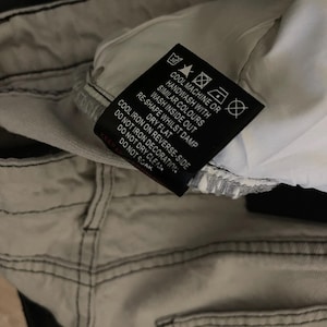 Puede incluir: Primer plano de unos vaqueros de mezclilla gris claro con costuras negras. Una etiqueta negra con texto blanco muestra las instrucciones de lavado, incluyendo "Lavar a m&aacute;quina en fr&iacute;o o a mano" y "Secar en plano". Los vaqueros est&aacute;n doblados, mostrando el forro interior.