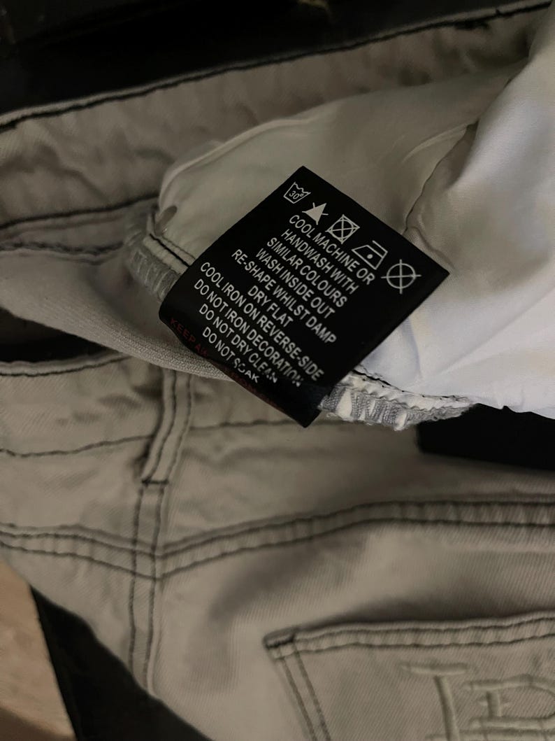 Puede incluir: Primer plano de unos vaqueros de mezclilla gris claro con costuras negras. Una etiqueta negra con texto blanco muestra las instrucciones de lavado, incluyendo "Lavar a m&aacute;quina en fr&iacute;o o a mano" y "Secar en plano". Los vaqueros est&aacute;n doblados, mostrando el forro interior.
