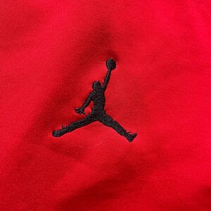 May include: Close-up of a red fabric with a black embroidered silhouette of a person with a basketball. The logo is a classic design associated with athletic apparel and footwear.