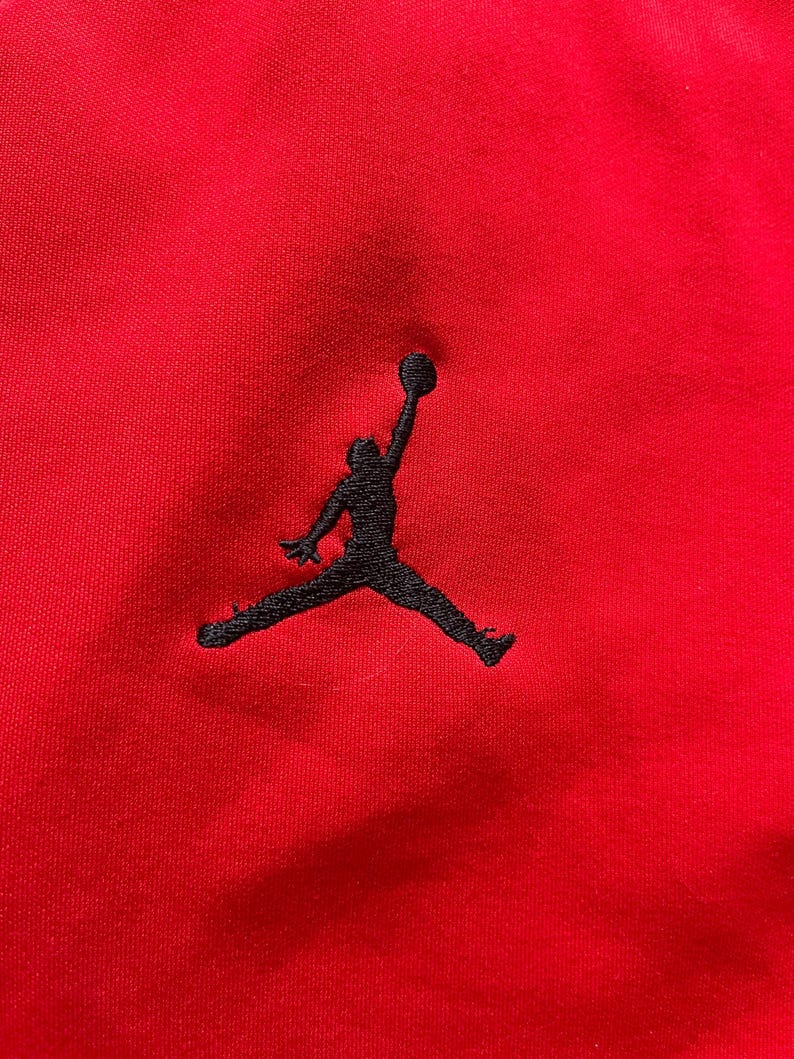 May include: Close-up of a red fabric with a black embroidered silhouette of a person with a basketball. The logo is a classic design associated with athletic apparel and footwear.