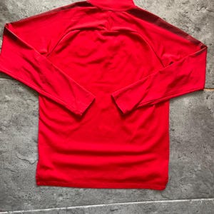 May include: A bright red long-sleeved athletic shirt with a mock neck collar. The shirt has dark red stripes on the shoulders and sleeves. The shirt is made of a smooth, possibly synthetic material.