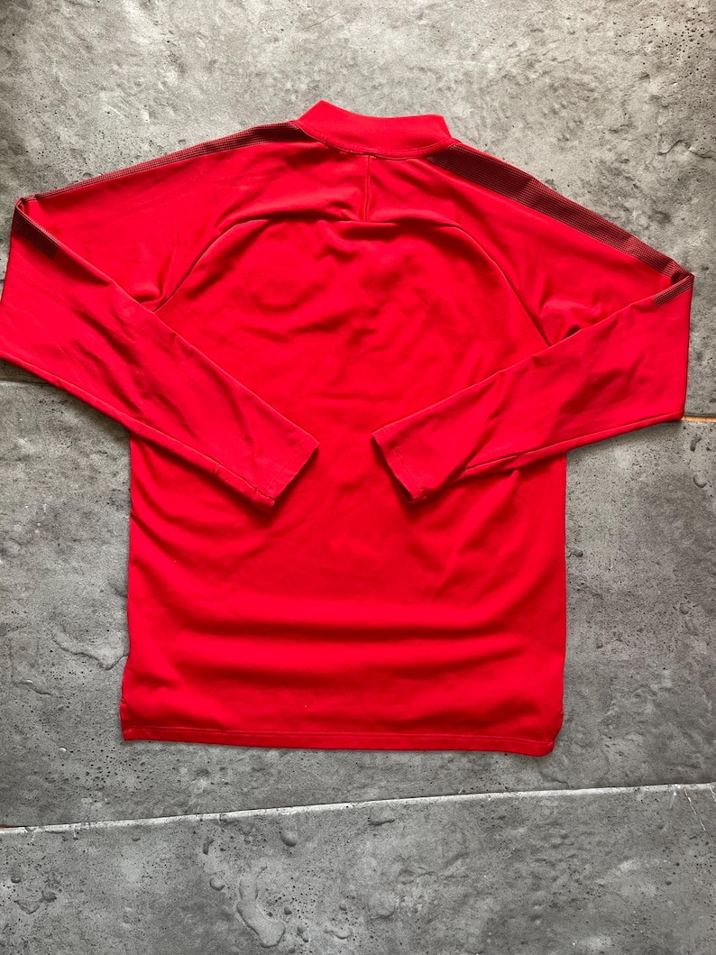 May include: A bright red long-sleeved athletic shirt with a mock neck collar. The shirt has dark red stripes on the shoulders and sleeves. The shirt is made of a smooth, possibly synthetic material.