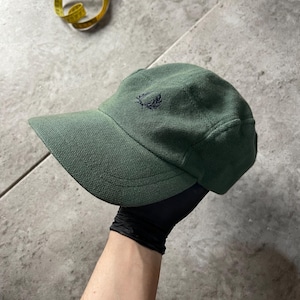 May include: A dark green baseball cap with a curved brim. The cap features a small, embroidered emblem on the front. The cap is held by a person wearing a black glove. A yellow measuring tape is in the background.