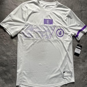 Può includere: Maglia sportiva bianca con dettagli viola. La maglia presenta la scritta "ULTRA" in viola sul petto, un logo circolare e un design geometrico. Le maniche hanno strisce viola. È presente un'etichetta Activision.