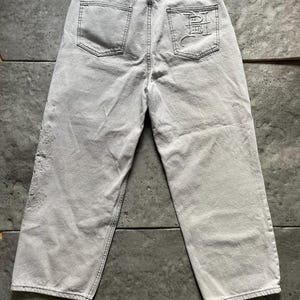 Puede incluir: Vaqueros de mezclilla gris claro con un dise&ntilde;o de pierna ancha. Los bolsillos traseros presentan un dise&ntilde;o bordado y la cintura tiene un parche de cuero negro. Los vaqueros tienen un aspecto descolorido y desgastado, con costuras visibles.