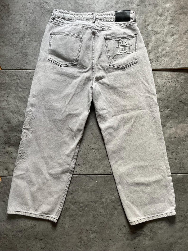 Puede incluir: Vaqueros de mezclilla gris claro con un dise&ntilde;o de pierna ancha. Los bolsillos traseros presentan un dise&ntilde;o bordado y la cintura tiene un parche de cuero negro. Los vaqueros tienen un aspecto descolorido y desgastado, con costuras visibles.