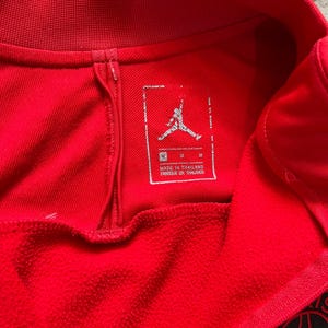 May include: Close-up of a red athletic jersey. The jersey features a tag with the Jumpman logo and text that reads "MADE IN THAILAND". The jersey has a ribbed collar and a zipper detail. A team logo is visible on the bottom right.