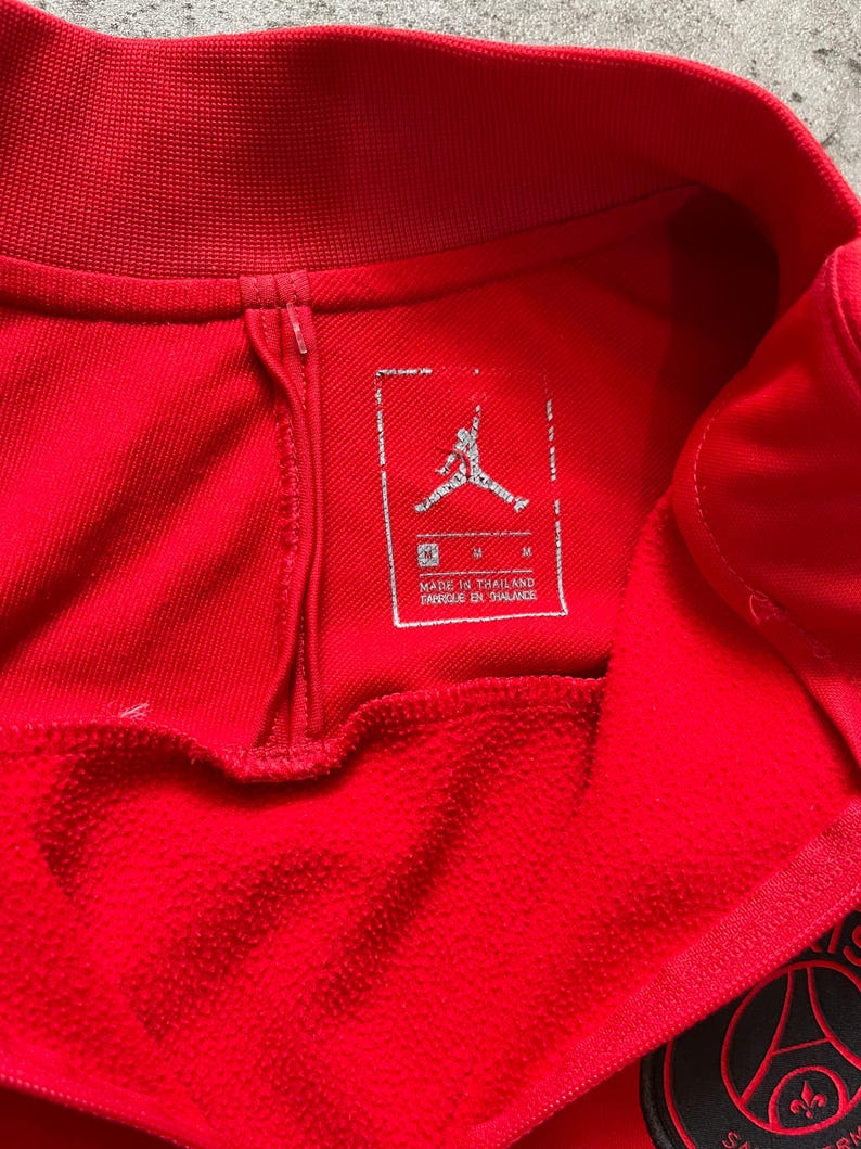 May include: Close-up of a red athletic jersey. The jersey features a tag with the Jumpman logo and text that reads "MADE IN THAILAND". The jersey has a ribbed collar and a zipper detail. A team logo is visible on the bottom right.