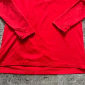 May include: Bright red long-sleeved athletic shirt with a quarter-zip neckline. Features a black Jordan logo on the upper left chest and a circular Paris logo on the upper right chest. The shirt has thumb holes at the cuffs.