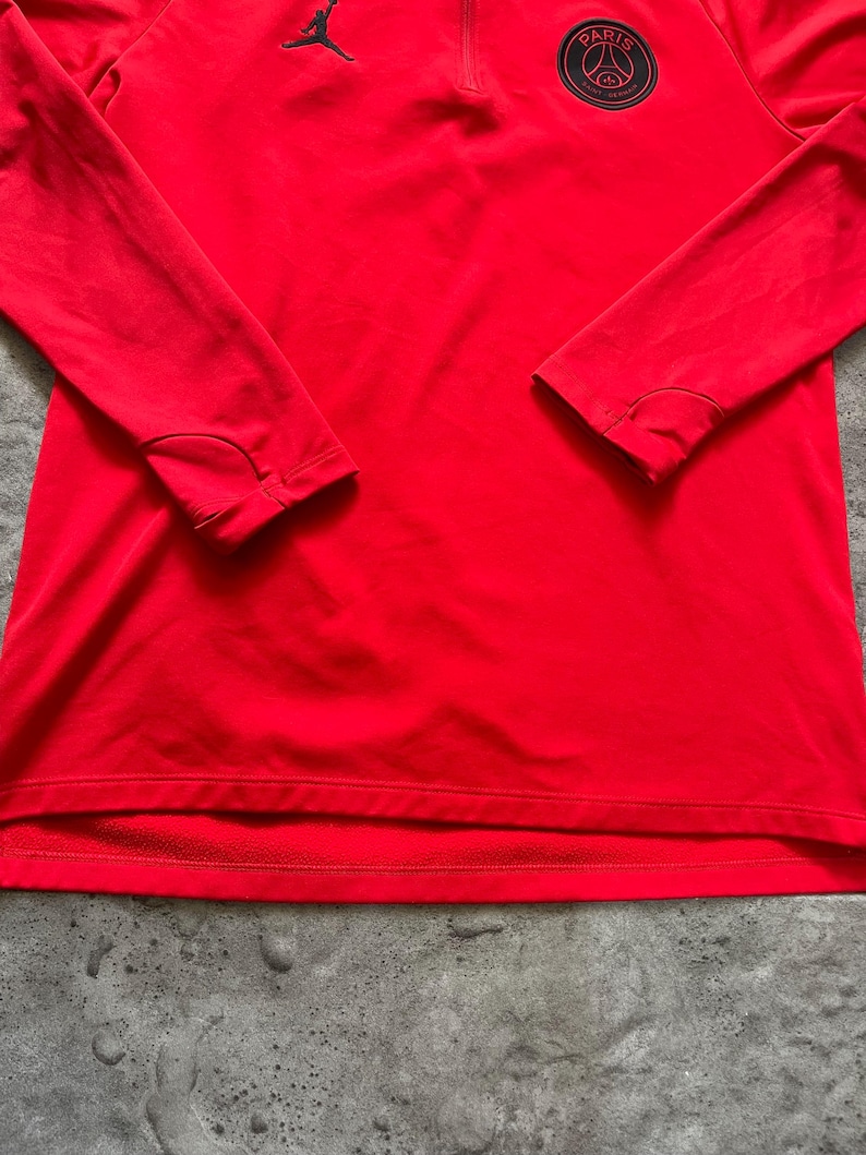 May include: Bright red long-sleeved athletic shirt with a quarter-zip neckline. Features a black Jordan logo on the upper left chest and a circular Paris logo on the upper right chest. The shirt has thumb holes at the cuffs.