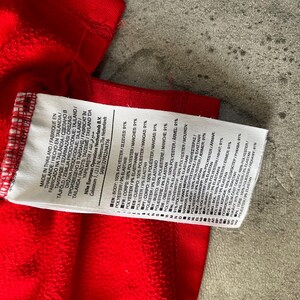May include: A close-up of a red garment with a white tag attached. The tag displays text in multiple languages, including English, French, and Spanish, detailing the fabric composition as 100% polyester and 9% elastane.